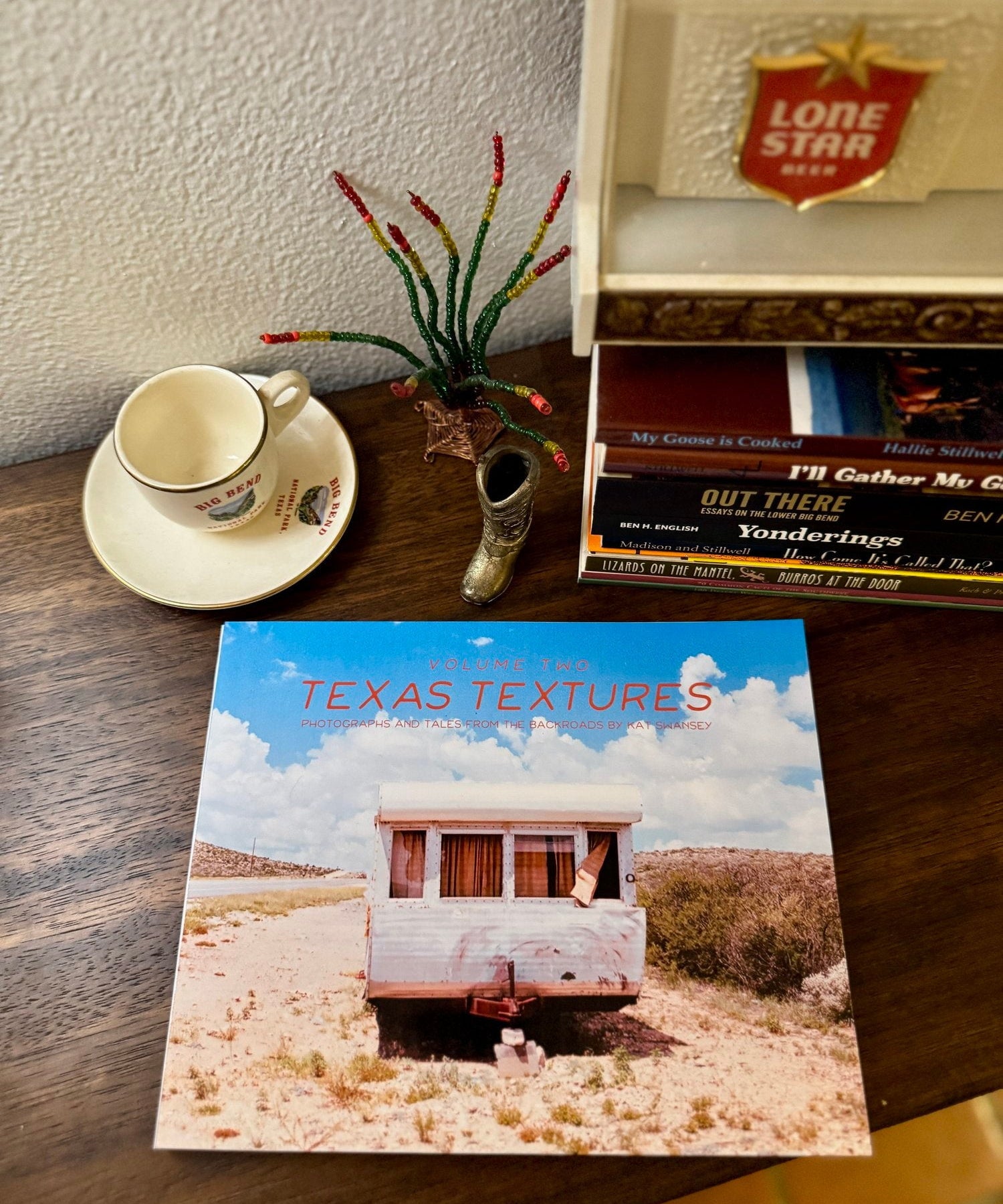 The "Texas Textures, Volume Two Book" by Kat Swansey rests on a table beside a teacup, decorative boot, and music CDs, its cover displaying a Kat Swansey photo of an abandoned Texas trailer.