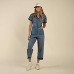 A woman stands against a beige background wearing The Dolly Jumpsuit by Sendero Provisions Co., paired with white sneakers, white socks, and a white cap featuring a small front graphic.