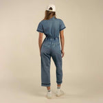 A person faces away, wearing The Dolly Jumpsuit by Sendero Provisions Co., paired with a white cap, striped socks, and white sneakers against a plain beige background.