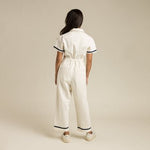 A person stands facing away from the camera, wearing The Dolly Jumpsuit White by Sendero Provisions Co. with dark trim and white sneakers, set against a plain beige background.