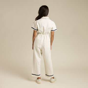 A person faces away against a beige background, wearing The Dolly Jumpsuit White by Sendero Provisions Co.—a white short-sleeve jumpsuit with black trim—paired with white sneakers.