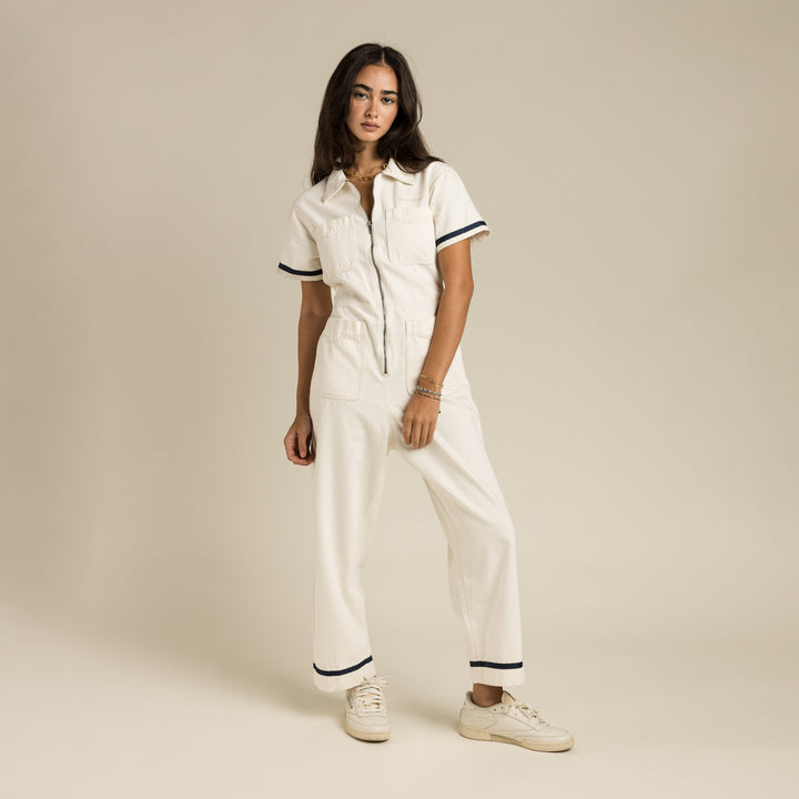 A woman stands against a plain beige background wearing the Sendero Provisions Co. Dolly Jumpsuit White, a crisp white women’s jumpsuit with short sleeves and black trim, paired with white sneakers.