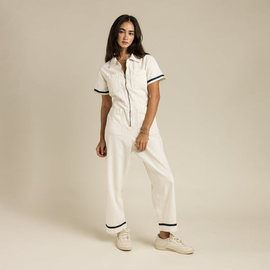 A woman stands against a plain beige background wearing the Sendero Provisions Co. Dolly Jumpsuit White, a crisp white women’s jumpsuit with short sleeves and black trim, paired with white sneakers.