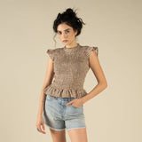 A woman with dark curly hair wears The Hattie Smock Top by Sendero Provisions Co. and light blue denim shorts while standing against a plain beige background.