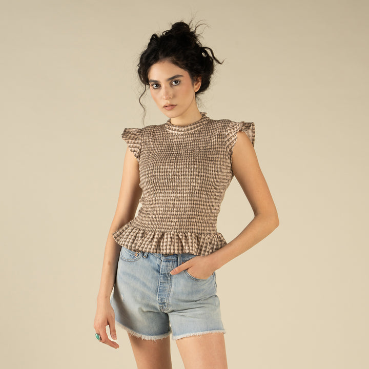 A woman with dark curly hair wears The Hattie Smock Top by Sendero Provisions Co. and light blue denim shorts while standing against a plain beige background.