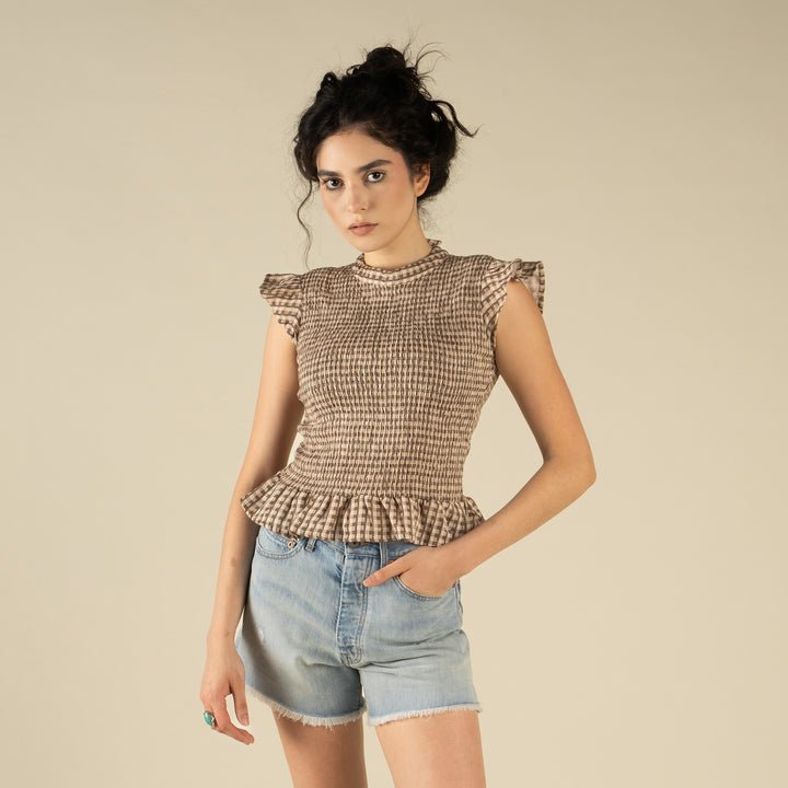 A woman with dark curly hair wears The Hattie Smock Top by Sendero Provisions Co. and light blue denim shorts while standing against a plain beige background.