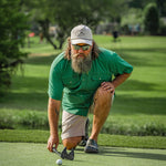 A man in The Magnolia | Poncho by Poncho kneels on a golf course, placing a golf ball on the green with a putter in hand.