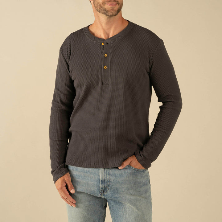 A man wearing The Maverick Henley by Sendero Provisions Co.—a long-sleeve, dark gray waffle knit shirt with three buttons—and light blue jeans stands against a plain beige background.