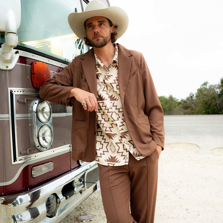 A man in a brown suit, The Padre Shirt | Chama Chevron by Sendero Provisions Co., and a white cowboy hat leans against a maroon bus on a dirt roadside.