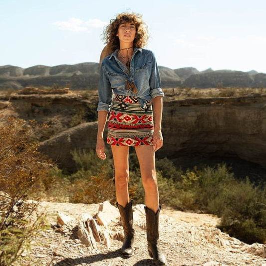 A woman stands on rocky terrain under a clear sky, wearing the Raquel Skirt by Sendero Provisions Co. with a denim shirt and cowboy boots against dry hills and brush in the background.