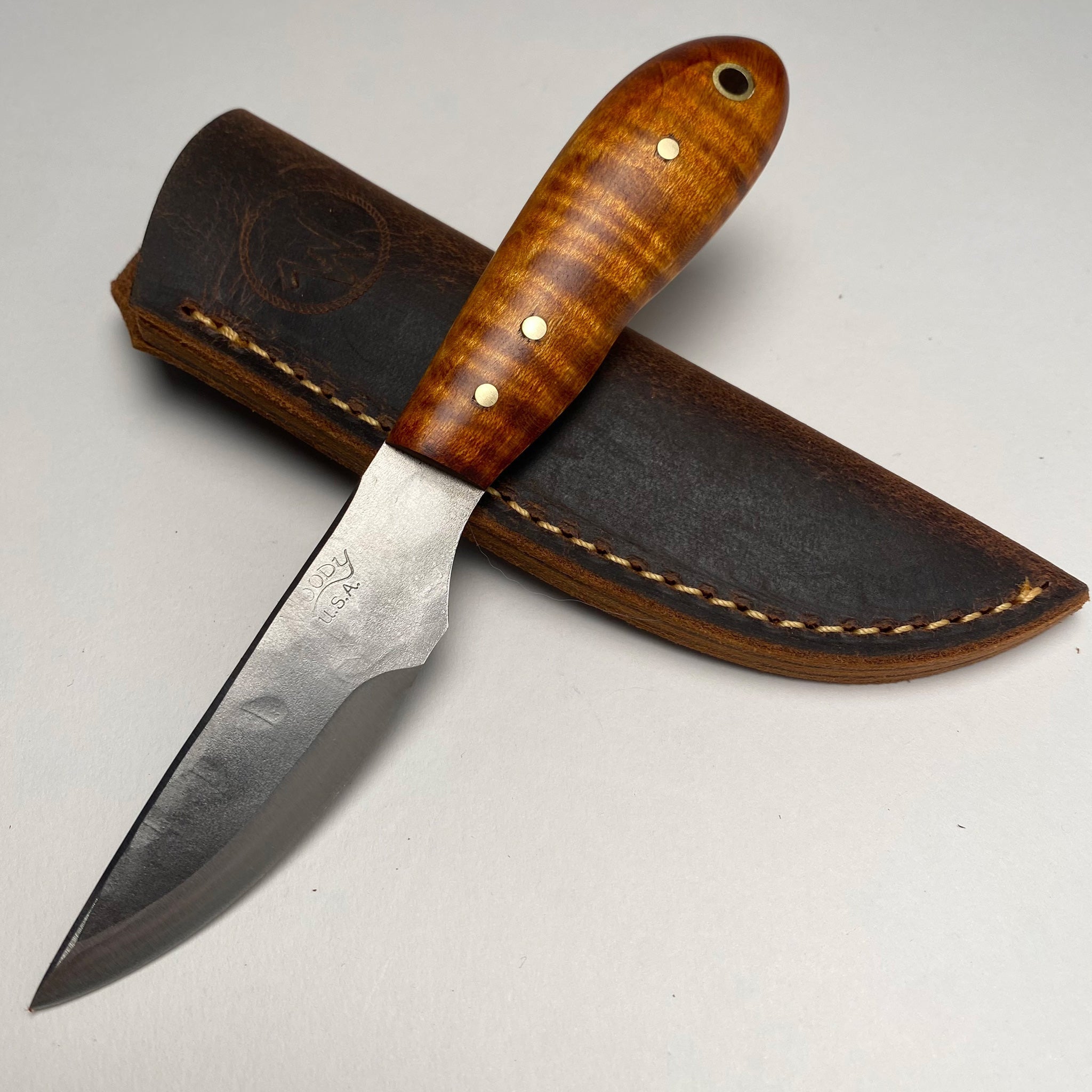 The Sidekick Long Clip by Woody Brand Knives features a handmade high-carbon tool steel fixed blade, a polished wood handle with brass rivets, and comes displayed on a brown leather sheath.