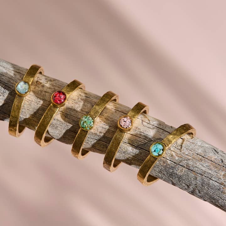 The Stacker Ring by cameoko features five gold-plated stackable rings with colorful round gemstones, displayed on a weathered wooden branch against a soft pink background.