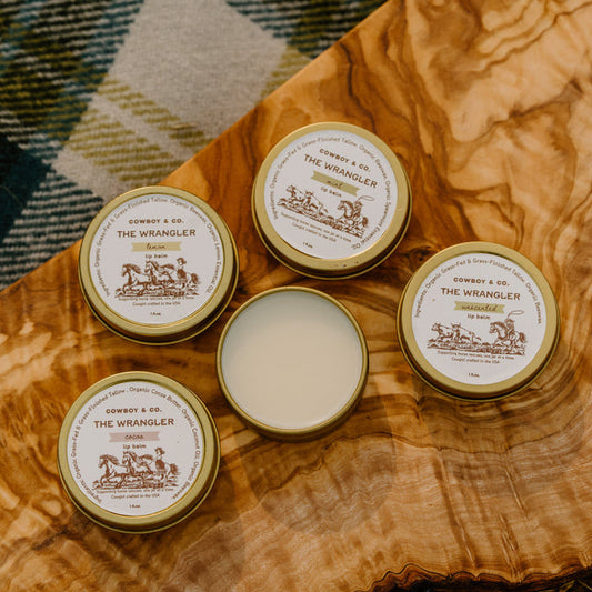 Four tins of The Wrangler Tallow Lip Balm by Cowboy & Co., one open to show its smooth contents, are displayed on a wooden surface with plaid fabric partially visible.