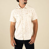 A man wearing The Wyatt Pearl Snap Dustbloom by Sendero Provisions Co.—a cream western shirt with a subtle floral pattern and pearl snaps—paired with dark jeans, stands against a plain beige background.