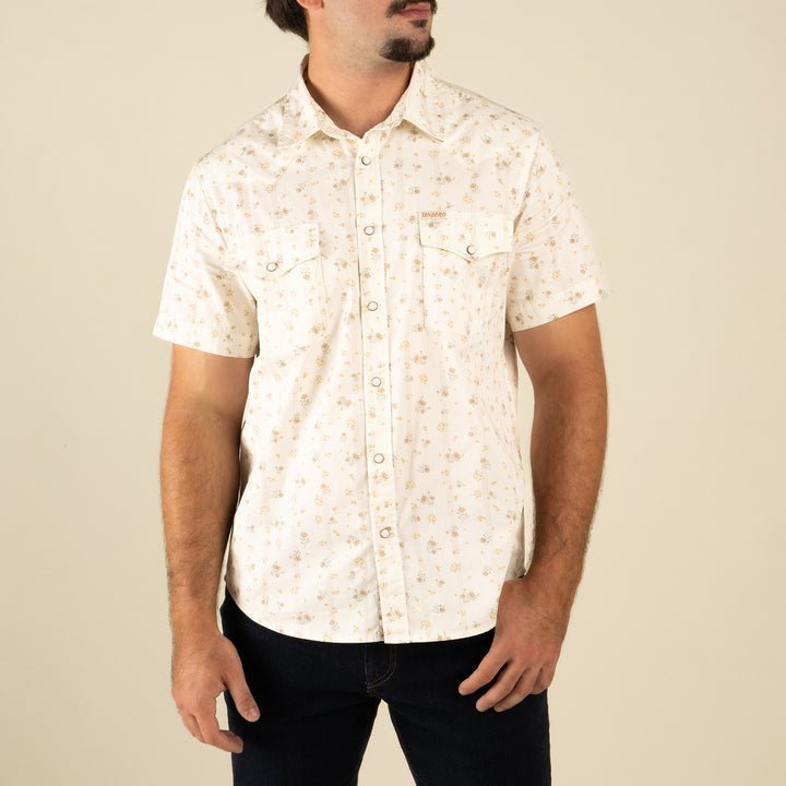 A man wearing The Wyatt Pearl Snap Dustbloom by Sendero Provisions Co.—a cream western shirt with a subtle floral pattern and pearl snaps—paired with dark jeans, stands against a plain beige background.