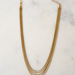 The Thelma Layered Snake Chain by Peter and June features four 18k gold plated chains, a lobster clasp, and an adjustable chain, elegantly displayed on a white marble surface.