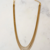The Thelma Layered Snake Chain by Peter and June features four 18k gold plated chains, a lobster clasp, and an adjustable chain, elegantly displayed on a white marble surface.