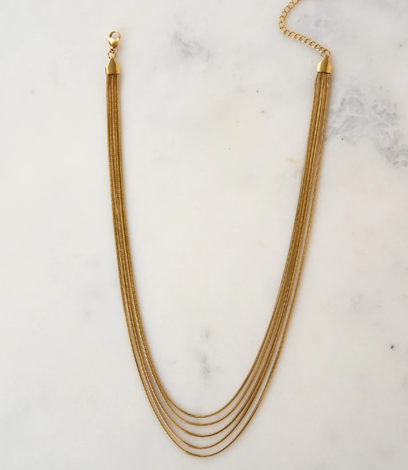 The Thelma Layered Snake Chain by Peter and June features four 18k gold plated chains, a lobster clasp, and an adjustable chain, elegantly displayed on a white marble surface.