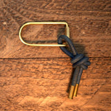 The Thistle Keychain by Tres Cuervos features an oval brass ring with a knotted navy blue leather tassel and brass tips, displayed on a textured wooden surface.