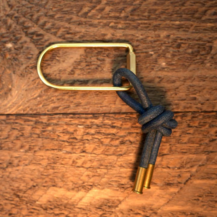 The Thistle Keychain by Tres Cuervos features a brass oval screwgate keyring attached to a looped navy blue leather cord with brass tips, displayed on a wooden surface.