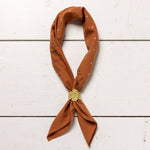 A rust-colored bandana with white and black geometric patterns is styled with a gold shell pin and the Jenni Earle Thunderbird Bandana Slide, displayed flat on a white wooden surface for an authentic western look.