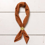 A rust-colored bandana with white and black geometric patterns is styled with a gold shell pin and the Jenni Earle Thunderbird Bandana Slide, displayed flat on a white wooden surface for an authentic western look.