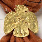 The Thunderbird Bandana Slide by Jenni Earle features a brass thunderbird holding arrows, fastened to brown patterned fabric for a bold western look.