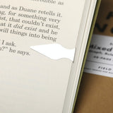 A precision-cut steel Book Dart from the Field Notes Tin-O-Darts set marks a page in an open book, with a Field Notes notebook partially visible nearby.