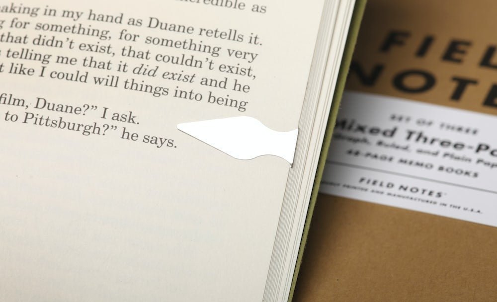 A precision-cut steel Book Dart from the Field Notes Tin-O-Darts set marks a page in an open book, with a Field Notes notebook partially visible nearby.