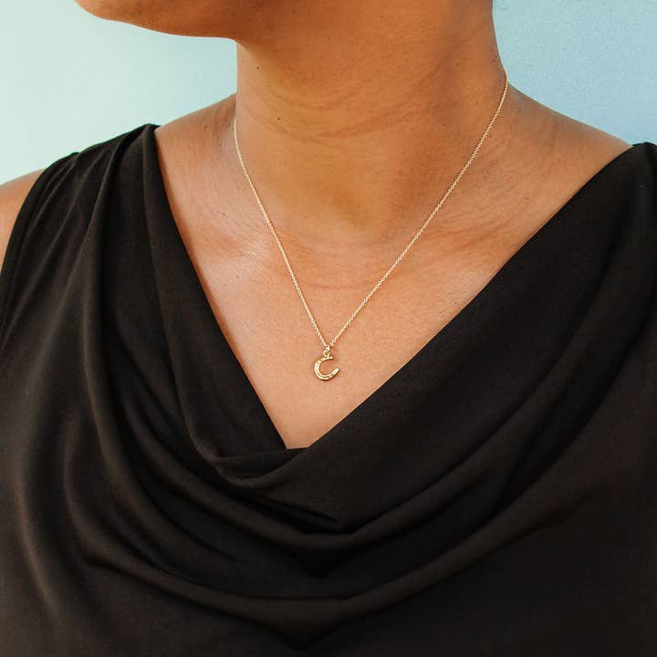 A person wearing a black draped top and the Tiny Gold Horseshoe Necklace by Ornamental Things is shown from shoulders to chest against a light blue background, showcasing an elegant minimalist jewelry style.