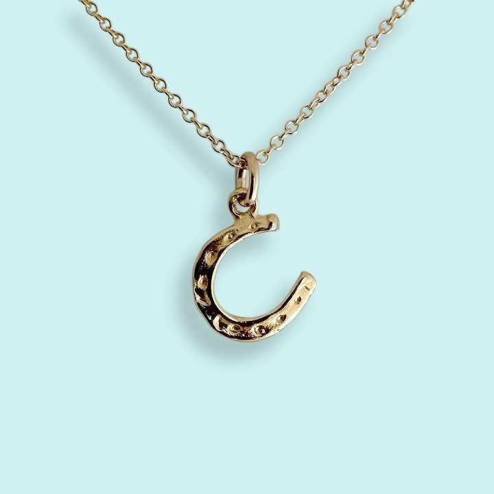 Tiny Gold Horseshoe Necklace by Ornamental Things: a 14k gold-plated chain with a horseshoe pendant, shown on a light blue background. This good luck necklace brings an elegant accent to your style.