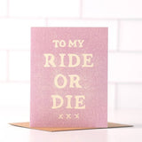 The "To My Ride Or Die" card by Daydream Prints features bold white lettering on pink, displayed on a brown envelope with a white tiled backdrop.