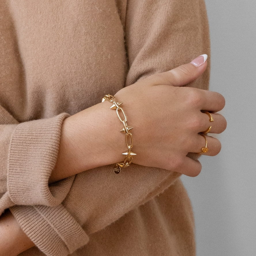 A person in a beige sweater wears the Larissa Loden Toom Spike Chain Bracelet—14k gold plated with a toggle clasp—and two gold rings on their left hand, arms crossed against a neutral background.