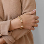 A person in a beige sweater wears the Larissa Loden Toom Spike Chain Bracelet—14k gold plated with a toggle clasp—and two gold rings on their left hand, arms crossed against a neutral background.