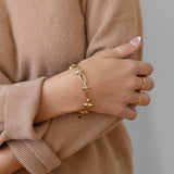 A person in a beige sweater wears the Larissa Loden Toom Spike Chain Bracelet—14k gold plated with a toggle clasp—and two gold rings on their left hand, arms crossed against a neutral background.