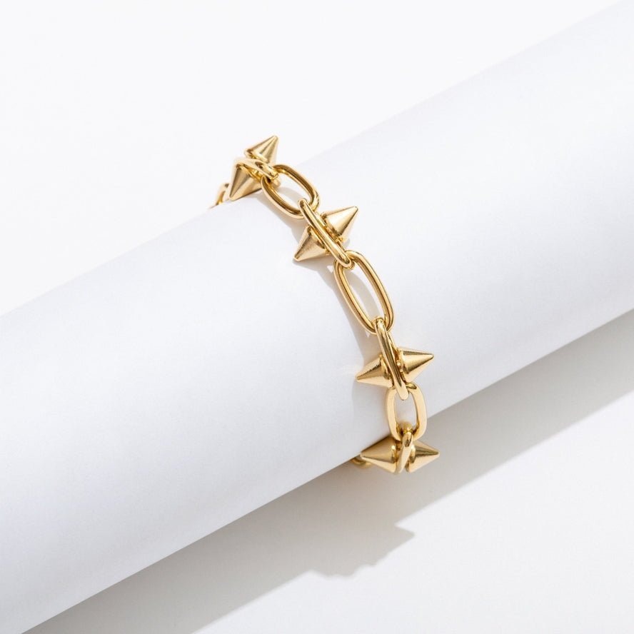 The Toom Spike Chain Bracelet by Larissa Loden features 14k gold plating, elongated oval links with spike accents, and a secure toggle clasp. Shown wrapped around a white cylindrical object against a white background.
