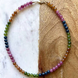 The Topanga Necklace by Jessica Matrasko Jewelry features round, glossy jade beads in pink, orange, green, and blue gradients—ideal for layering with your favorite 14k gold filled or layered necklaces. Displayed on a marble and wood surface.
