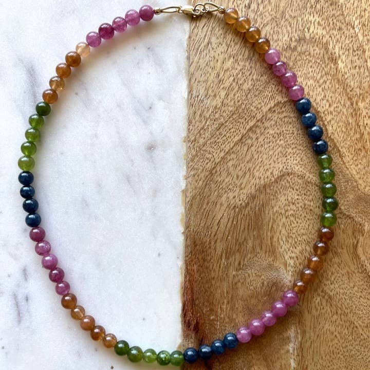 The Topanga Necklace by Jessica Matrasko Jewelry features round, glossy jade beads in pink, orange, green, and blue gradients—ideal for layering with your favorite 14k gold filled or layered necklaces. Displayed on a marble and wood surface.
