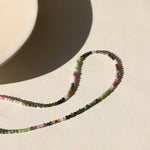 The Tourmaline Beaded Necklace by Token Jewelry, featuring hypoallergenic multicolored tourmaline beads, is displayed on a light surface beside the curved edge of a round white plate.