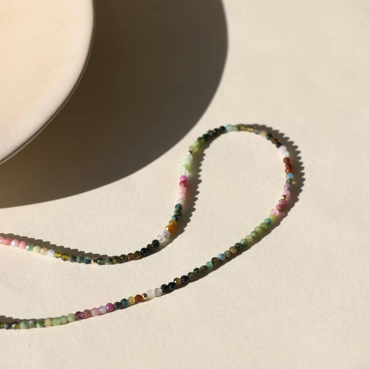 The Tourmaline Beaded Necklace by Token Jewelry, featuring multicolored beads, rests on a light surface next to the edge of a round cream-colored dish.