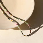 The Tourmaline Beaded Necklace by Token Jewelry, with multicolored tourmaline beads and a hypoallergenic finish, is draped over a round light ceramic dish on a cream background.