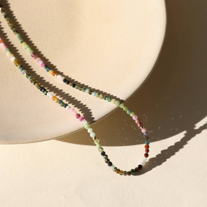 The Tourmaline Beaded Necklace by Token Jewelry features multicolored tourmaline beads, beautifully displayed on a light beige plate and surface, casting a subtle shadow.