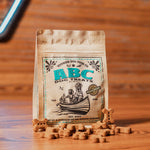 A brown bag of Voyager Dog Food Co. ABC Dog Treats | Voyager is displayed on a wooden surface, with small bone-shaped treats scattered in front.
