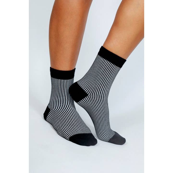 A person wearing Tailored Union's Trey Ankle Sock—black and white vertical stripes with solid black toes, heels, and cuffs—stands on a light background.