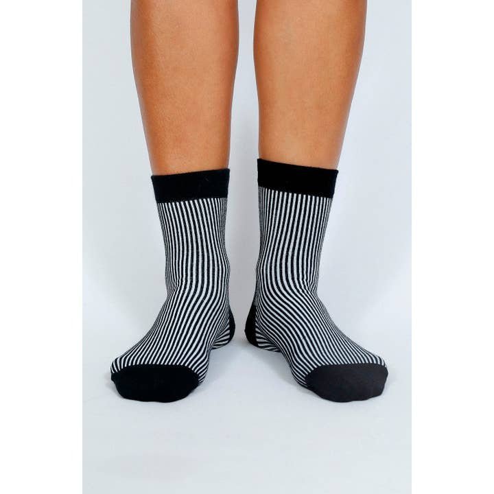 A person wearing Tailored Union Trey Ankle Socks—black and white vertically striped combed cotton with black toes, heels, and cuffs—stands on a white background.