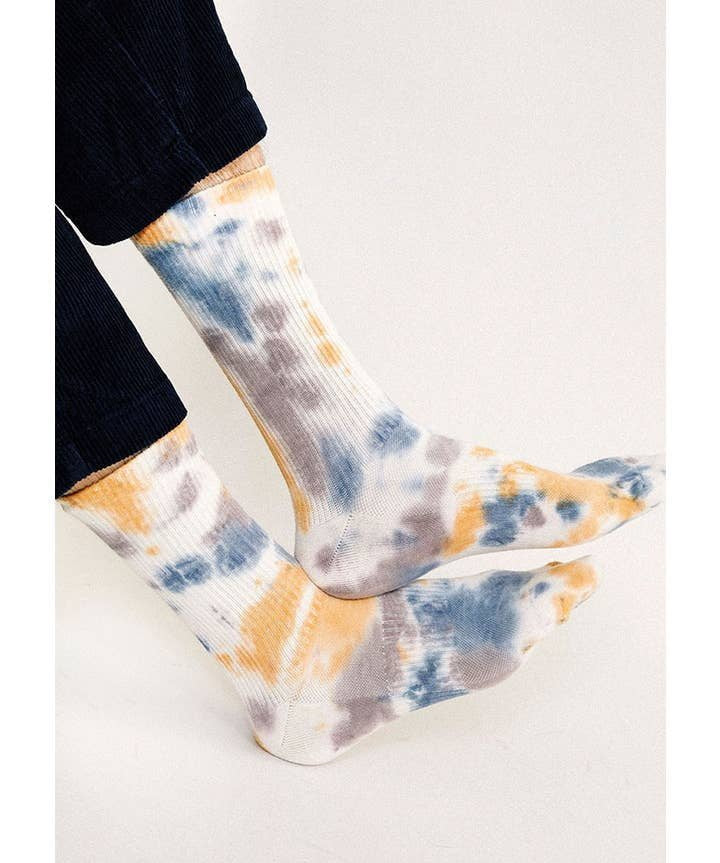 Person wearing Trippy Crew Socks by Tailored Union, featuring a colorful tie-dye design in blue, yellow, and gray, standing against a plain light background.