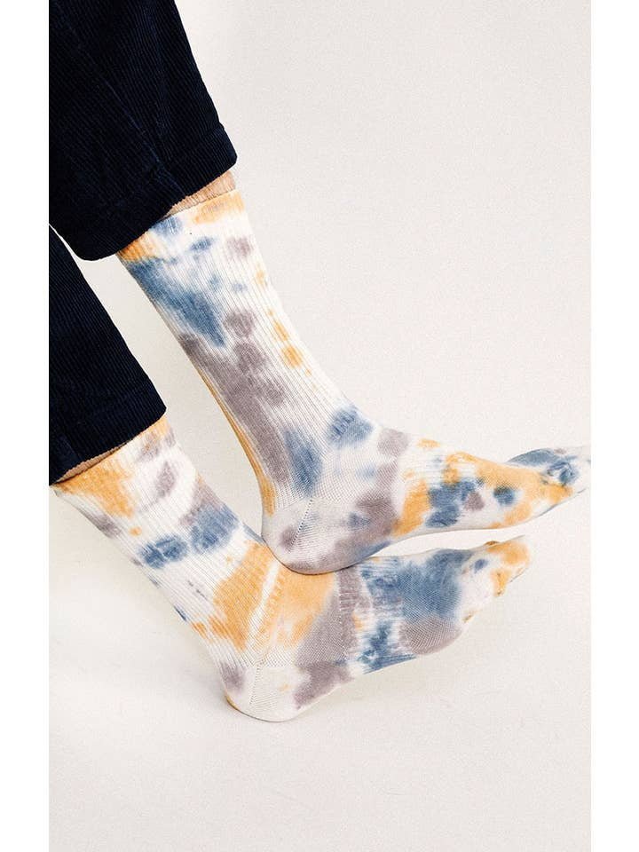 Person wearing Trippy Crew Socks by Tailored Union, featuring a colorful tie-dye design in blue, yellow, and gray, standing against a plain light background.