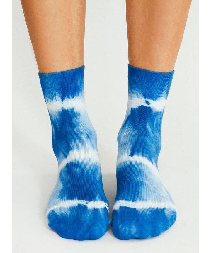 Wearing Tailored Union’s Trippy Tie Dye Ankle Sock-Blue, a pair of feet stand on a light surface, channeling fun festival vibes with their bold blue and white design.