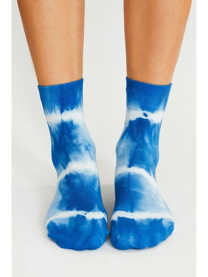 Wearing Tailored Union’s Trippy Tie Dye Ankle Sock-Blue, a pair of feet stand on a light surface, channeling fun festival vibes with their bold blue and white design.