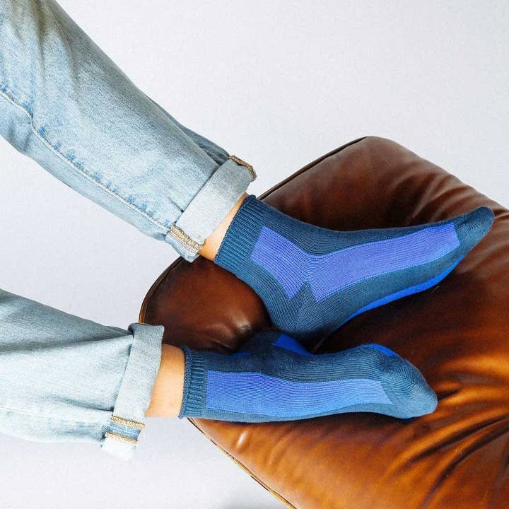 A person wearing Trois Striped Ankle Socks by Tailored Union with rolled-up jeans rests their feet on a brown leather ottoman against a light background.